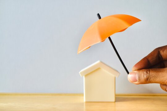 Hand holding a small orange umbrella over a model house model on a wooden table, symbolizing home insurance, security, and property protection.