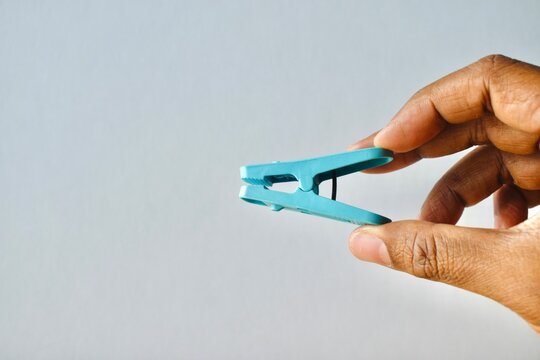 Hand holding a blue plastic clothes peg against a neutral light grey background, representing laundry accessories and pinch concepts.