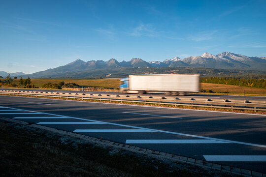 Motion blurred cargo truck driving on the D1 motorway beneath the High Tatras in Slovakia. Road freight transport and logistics against snow dusted mountain peaks and autumn countryside.