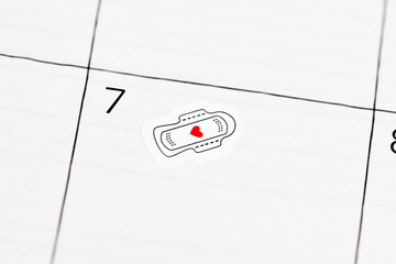 Close up of calendar with sticker showing sanitary pad and red heart. Reminder for tracking menstrual cycle and period start © Firn