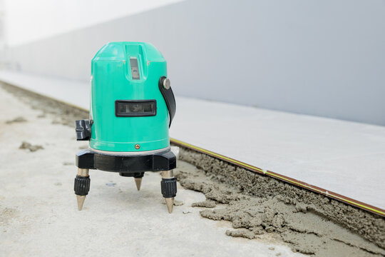 Green laser level tool set on a tripod, aligning ceramic tiles on a white and gray floor. Essential equipment for accurate tiling work.