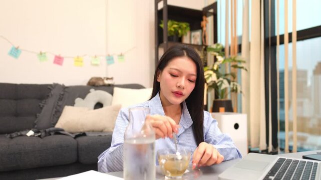 Young Asian Woman Stirring Tea While Working from Home on Laptop