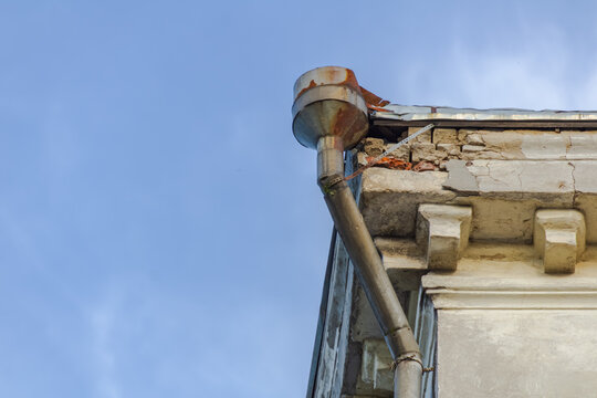 Old rusty metal downspout attached to a damaged brick cornice on an old building corner against a blue sky, weathered architectural detail, urban decay and building maintenance concept