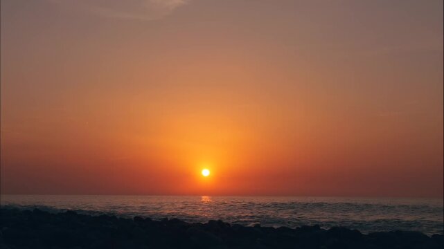 Sunset Timelapse Over Calm Sea with Sun Sinking Below Horizon, Glowing Orange Sky at Golden Hour Fading into Dusk Above Rocky Shore and Gentle Waves.