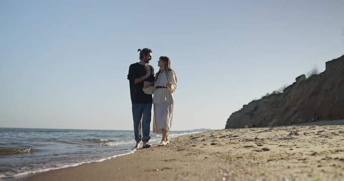 Two people walking hand in hand along wet sandy shoreline by steep sand cliff with gentle waves and clear blue sky, barefoot stroll showing relaxed intimate connection