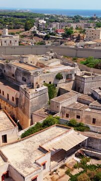 An aerial view shows a weathered masonry complex with flat roofs and courtyards, surrounded by colorful buildings, trees and fields on Favignana, with the sea beyond.