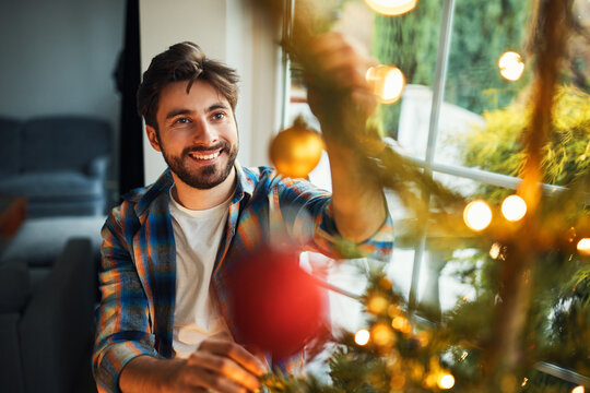 Smiling young man decorating christmas tree with ornaments at home