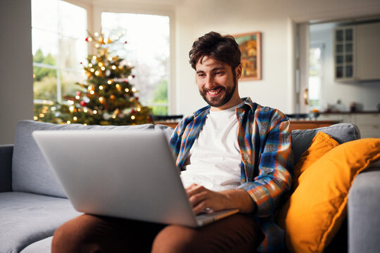 Smiling man using laptop on sofa near christmas tree at home