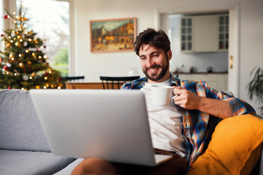 Smiling man holding coffee cup and using laptop in living room at christmas