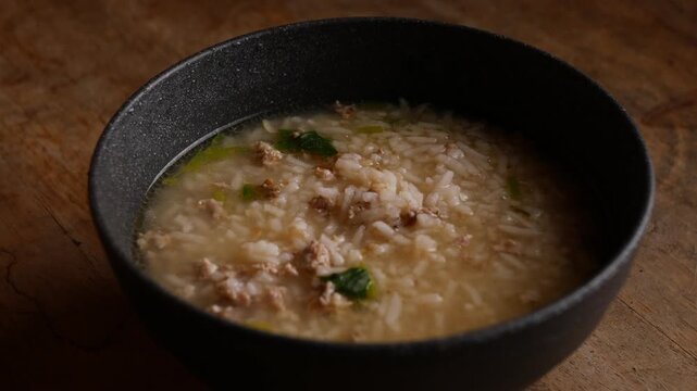 Thai rice soup with minced pork, bok choy and green onion in clear savory broth served in a dark ceramic bowl on a rustic wooden table, comforting breakfast dish