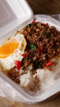 Thai basil stir-fried minced pork with crispy fried egg over steamed rice in a white foam takeaway box with plastic spoon, street food lunch on a rustic wooden table