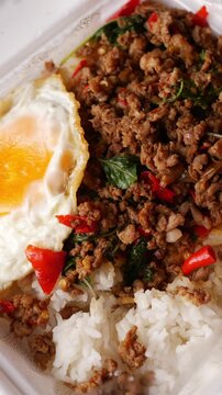 Thai basil stir-fried minced pork with crispy fried egg over steamed rice in a white foam takeaway box with plastic spoon, street food lunch on a rustic wooden table