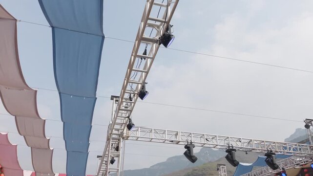 Outdoor Stage Truss with Colorful Lighting Fixtures Beneath Fabric Canopy, Mountain Backdrop and Blue Sky for Open Air Event Setting Under Construction