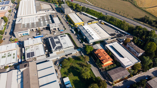 Aerial view of an industrial zone featuring warehouses with solar panels, flat roofs, and surrounding green spaces in Opera, Lombardy, Italy.