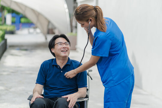 Smiling Asian female nurse using a stethoscope to examine a senior male patient in a wheelchair.