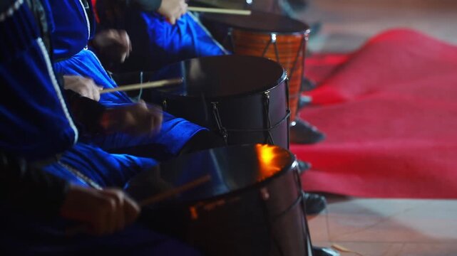 Musicians dressed in blue uniforms beat traditional drums rhythmically with wooden sticks while sitting down in a row indoors on a red floor