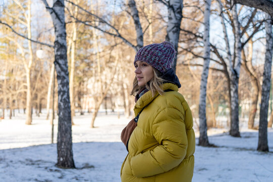 Caucasian pregnant woman in yellow coat taking contemplative walk through sunlit snowy park with bare trees, purple beanie, hand on belly, soft backlight and serene mood