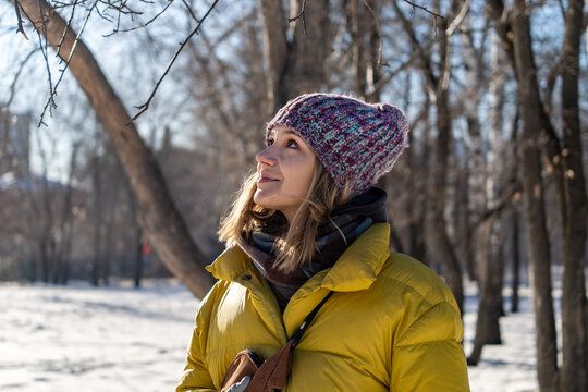 Young woman wearing a yellow jacket and hat walking through a snowy city park, smiling and enjoying a bright winter day. Waist-up view capturing her cheerful mood and pleasant seasonal atmosphere.