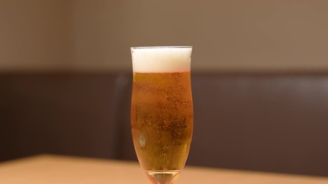 A glass of amber lager beer in tulip glass is placed on wooden table, with foam on top and bubbles floating up. Drink object footage, close-up with selective focus.