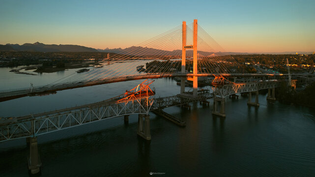 Aerial view of the SkyBridge and Pattullo Bridge spanning the Fraser River during golden hour with mountains in the background in New Westminster, British Columbia, Canada.