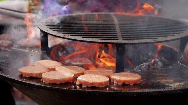 Pork patties cook on outdoor griddle over open flames, sending smoke above round iron cooking surface. Fresh meat sizzles beside charcoal at lively outdoor food event, cooking on hot iron.