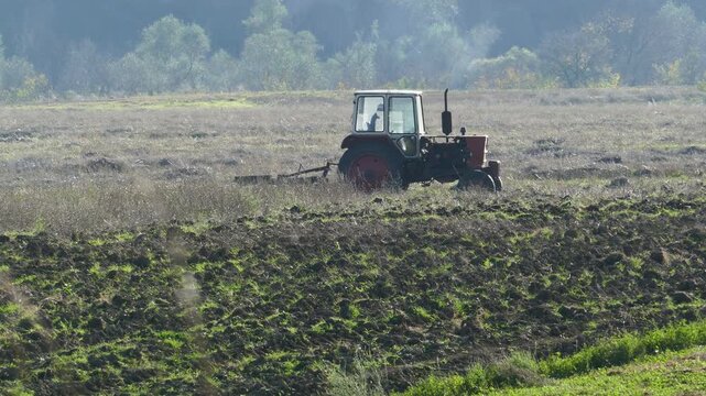 Distant shot of an old Soviet-era vintage tractor maneuvering near an overgrown fallow field, historic Eastern Bloc agriculture machinery working by plowed land and wild grass in Bulgaria
