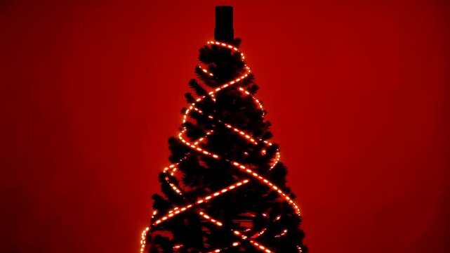 Christmas tree wrapped in glowing string lights as illumination pattern shifts across branches against a dramatic red background, festive holiday decoration in studio