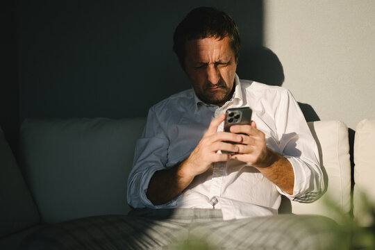 Concerned middle-aged man sitting on a sofa and looking at his smartphone in strong sunlight at balcony. Digital communication, stressful news, uncertainty and modern lifestyle concept.
