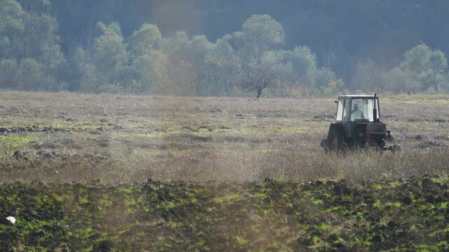 Distant shot of an old Soviet-era vintage tractor maneuvering in a field with an unrecognizable driver, historic Eastern Bloc agriculture machinery working on a farm plantation in Bulgaria
