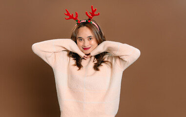 Asian woman wearing reindeer headband smiling and posing with hands under chin on brown studio background with copy space.  © Ryan
