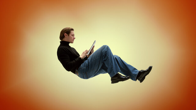 Attentive young adult browsing tablet while levitating against orange background. Mobile apps, digital publishing concept.