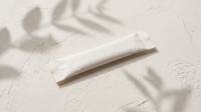 Single blank white paper stick sachet on textured white plaster surface, delicate blurred plant shadows, monochromatic styling, product design. Mock-up. Close-up.