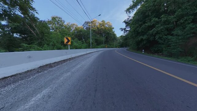 Winding asphalt road through dense green forest, featuring a concrete roadside barrier, yellow curve warning signs, overhead power lines and a vehicle in the distance beneath a blue sky.