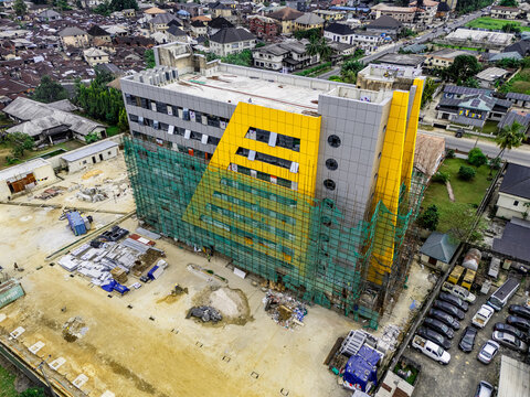Aerial view of the NDDC CMS building under construction, featuring a yellow and grey facade with scaffolding, in an urban area with a parking lot in Port Harcourt, Rivers, Nigeria.