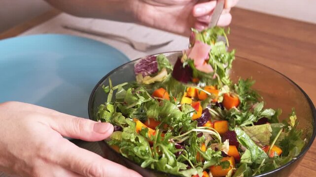 person serving fresh vegetable salad from glass bowl onto blue plate using fork during home meal, showcasing colorful ingredients, greens, roasted pumpkin, beetroot and careful healthy cooking