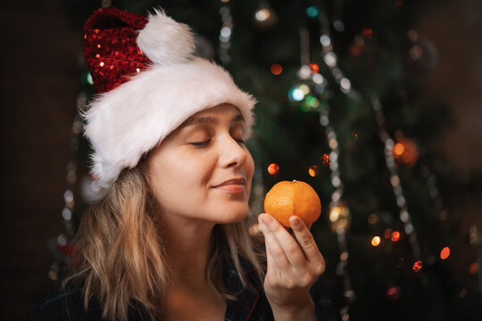 Girl in Santa hat smelling a mandarin with closed eyes, enjoying the citrus aroma. Christmas tree background, holiday sensory pleasure.