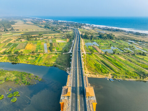 Aerial view of Kabanaran Bridge spanning a river, surrounded by lush fields, with the coastline and ocean in the background in Kecamatan Srandakan, Daerah Istimewa Yogya, Indonesia.
