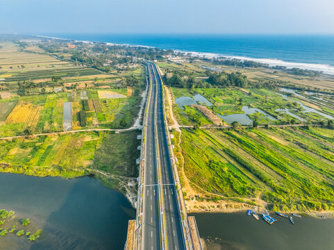 Aerial view of Kabanaran Bridge spanning the river, surrounded by green fields and wetlands, with the coastline in the distance in Kecamatan Srandakan, Daerah Istimewa Yogya, Indonesia.