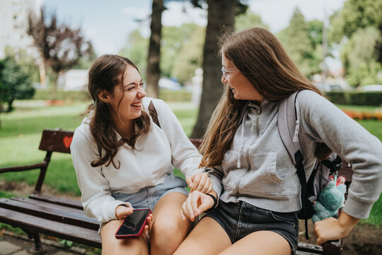 Two teenage friends laugh together on a park bench, holding a smartphone and backpacks. They share a casual, happy moment outdoors in a green city park, smiling and chatting.