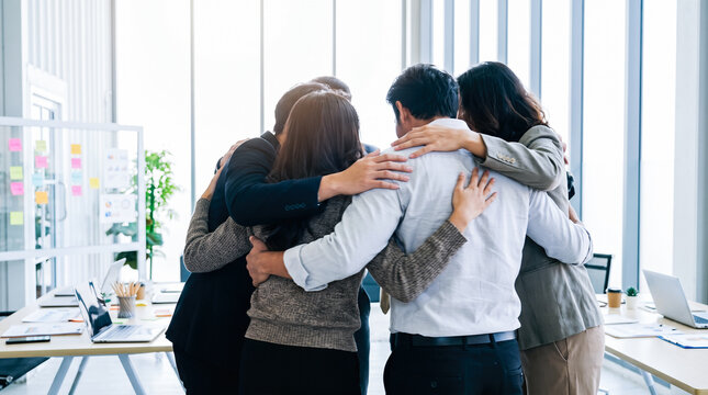 diverse team of business coworkers standing in tight supportive huddle embracing each other inside modern sunlit conference office room
