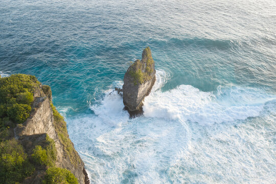 Aerial view of a dramatic sea stack rising from turquoise ocean waters with crashing white waves against the rugged cliffs of Nusa Penida, Bali, Indonesia.