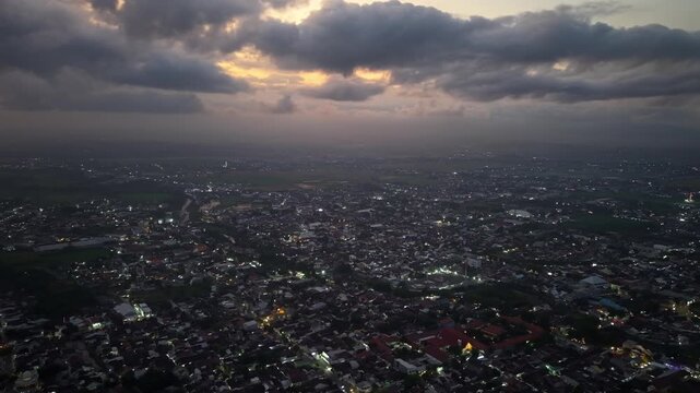 Sky darkens as city lights glow below. Clouds gather, casting shadows over urban sprawl at Lumajang