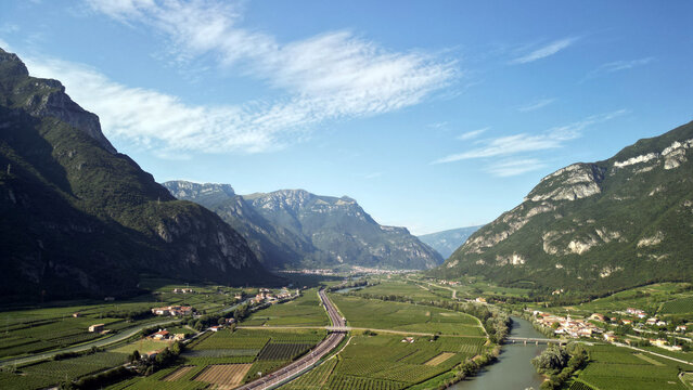 Aerial landscape of a green Alpine valley in Austria, featuring vineyards, a winding river, mountain slopes, rural settlements and transport infrastructure under a clear blue sky. 