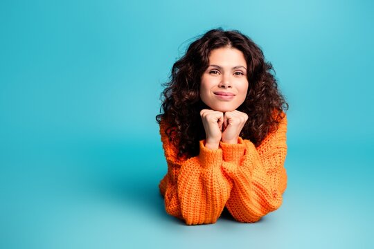 Smiling young woman in orange sweater lying on turquoise background, portrait with curly hair and relaxed thoughtful expression