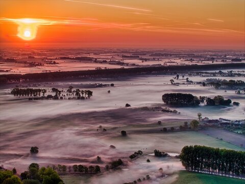 Aerial view of the misty landscape of Damme at sunrise, with golden light illuminating the fog-covered fields and rows of trees in Damme, Flanders, Belgium.