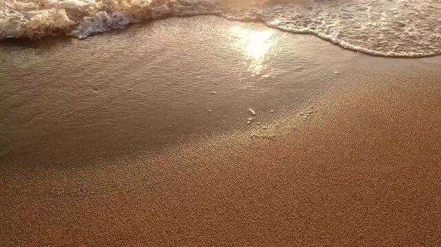 Close-up macro video of gentle golden ocean waves washing smoothly over wet sandy beach during sunset. Sparkling sunlight reflections on wet sand