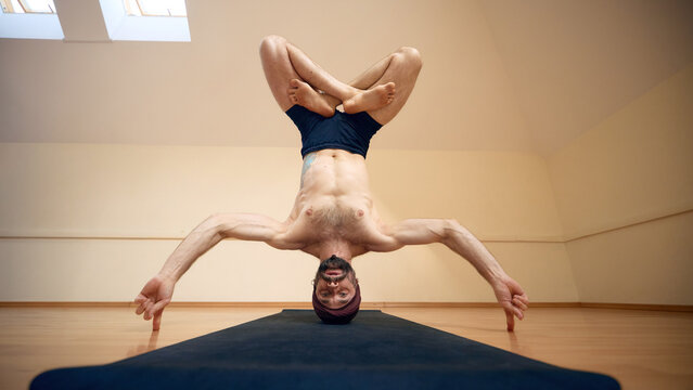 Focused man holding lotus headstand with arms extended sideways in yoga studio. Sports photography, advanced yoga events concept.