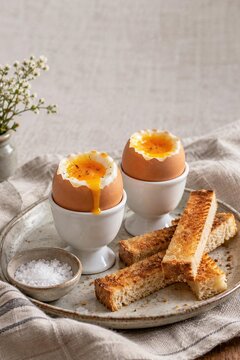 Two soft boiled eggs with runny yolks in white egg cups with toast soldiers, sea salt on ceramic plate with linen napkin