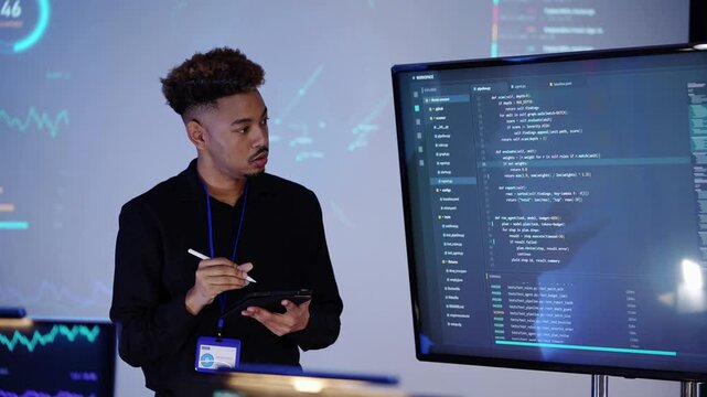 Young Black software engineer with curly blond-tipped hair writes on a tablet with a stylus, then points the pen at Python code on a large monitor in a dark blue AI office