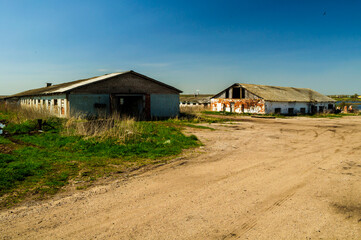 Abandoned industrial background with two old dilapidated barns featuring weathered brick walls and rusty corrugated roofs beside a dirt road © syntheticmessiah